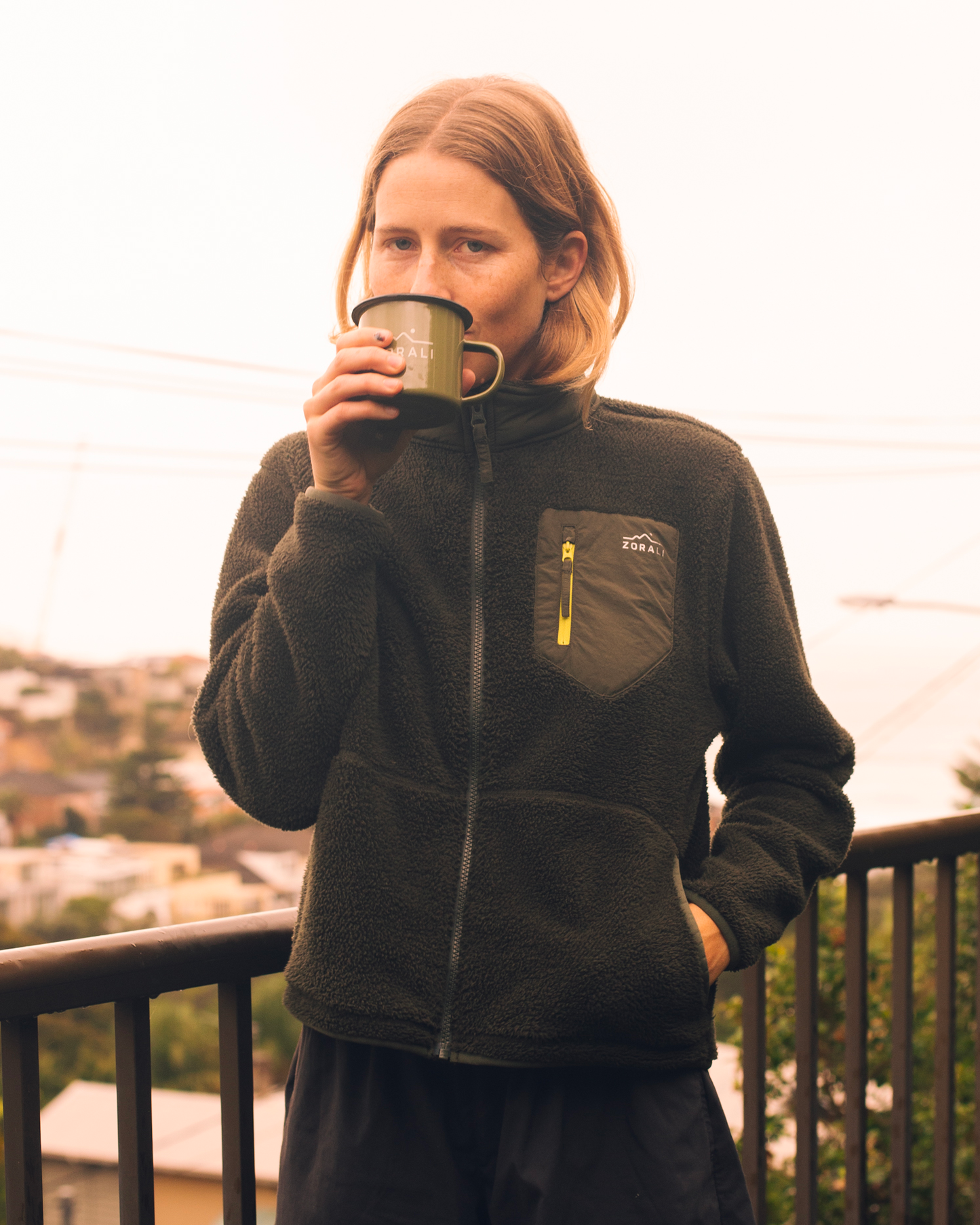 A woman wearing a dark olive green fleece zip-up jacket with a chest pocket and yellow zipper pull, holding a green enamel mug while standing on a balcony with a city view in the background.