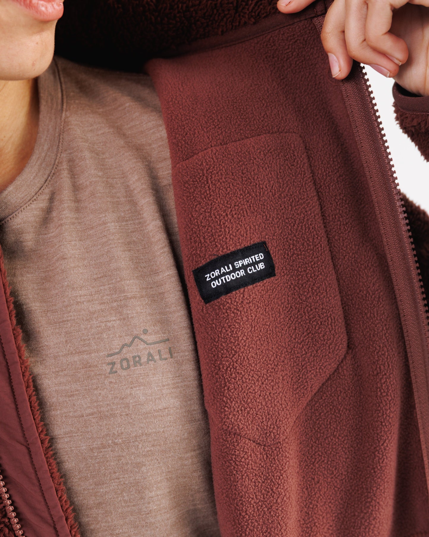 Close-up of a hand opening the front zip on a maroon sherpa fleece jacket, revealing the 'Zorali Spirited Outdoor Club' patch and tan t-shirt underneath.