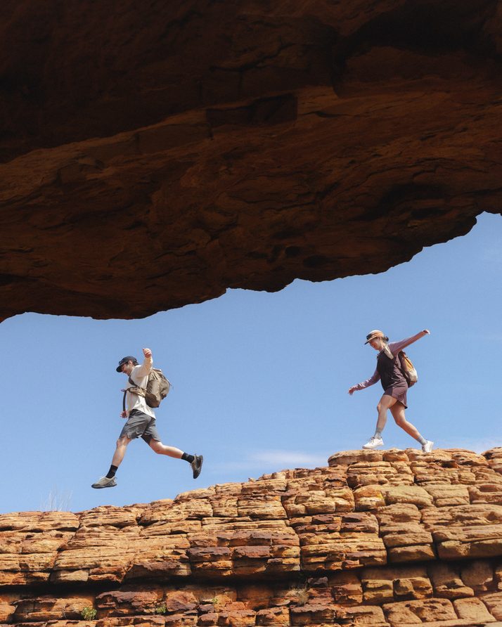 Two hikers leaping across a sandstone ridge under a rock arch at Kings Canyon