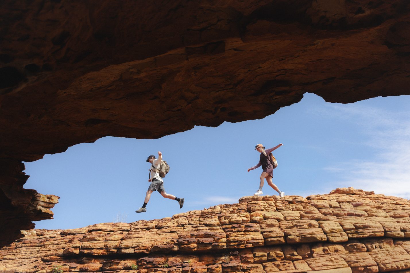 Two hikers leaping across a sandstone ridge under a rock arch at Kings Canyon