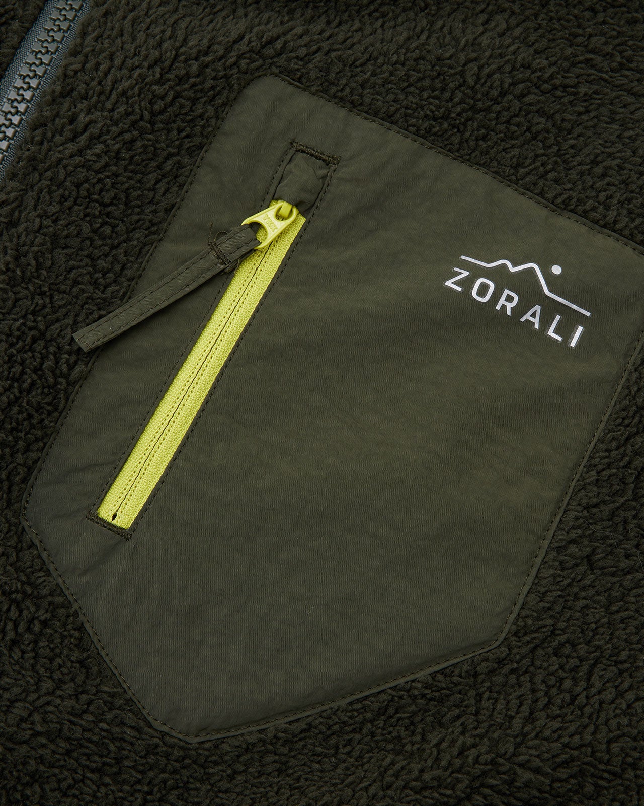 Close-up of the olive green chest pocket with yellow zipper pull and white Zorali mountain logo on a dark green sherpa fleece jacket.