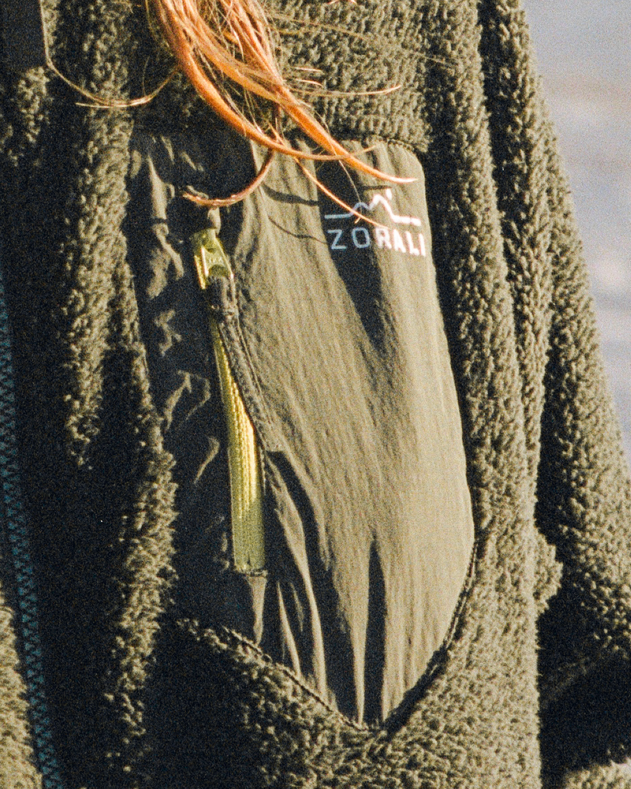Close-up of a dark olive green fleece jacket with a zippered pocket featuring a white Zorali logo and a gold zipper pull, worn by a person with orange hair visible.