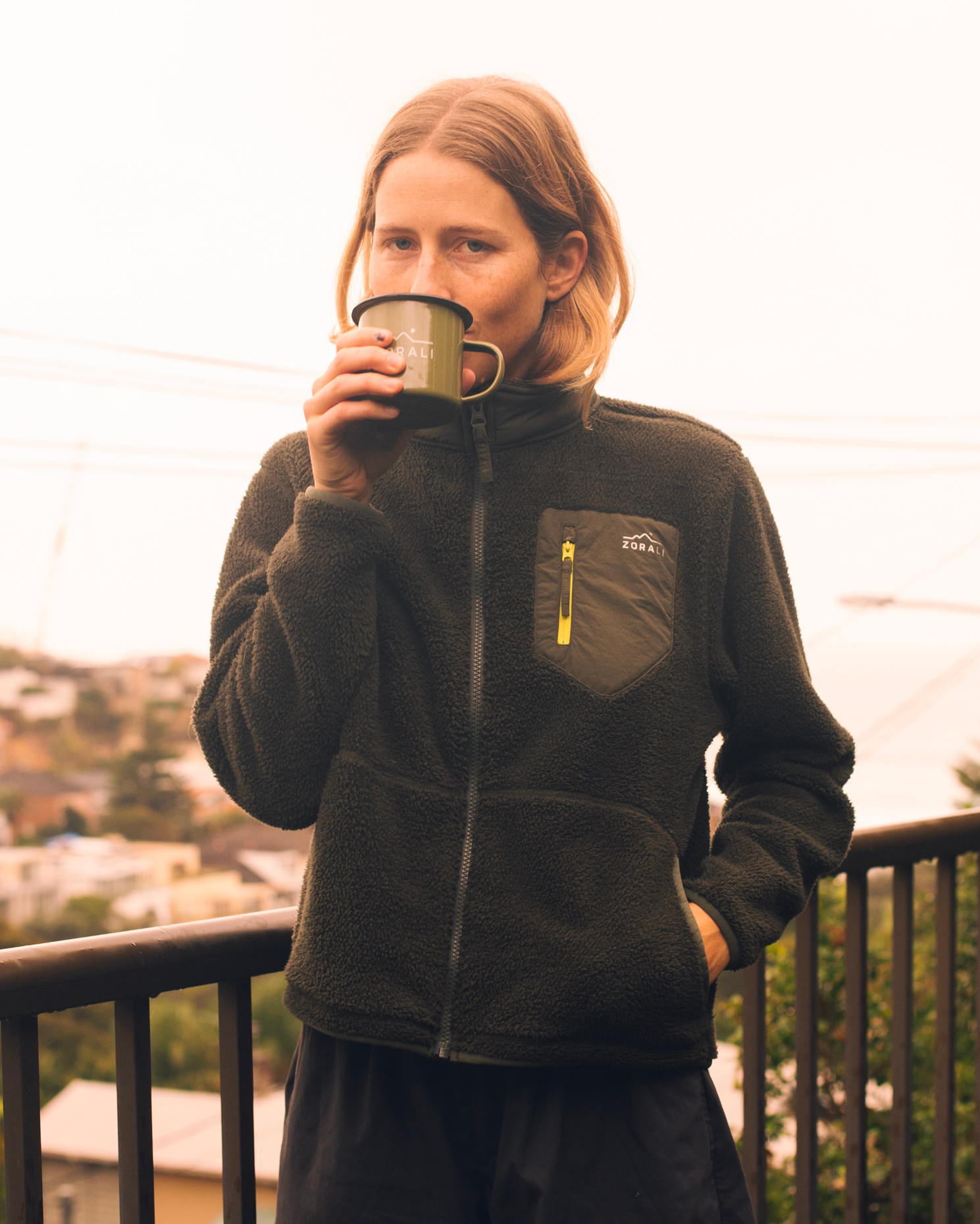 A woman wearing a dark olive green fleece zip-up jacket with a chest pocket and yellow zipper pull, holding a green enamel mug while standing on a balcony with a city view in the background.