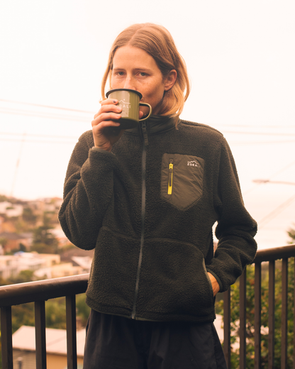 A woman wearing a dark olive green fleece zip-up jacket with a chest pocket and yellow zipper pull, holding a green enamel mug while standing on a balcony with a city view in the background.