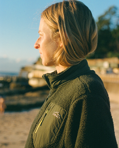 A woman in profile wearing an olive green fleece jacket with a visible Zorali logo on the chest pocket, photographed outdoors on a sandy beach with warm sunlight and a blurred background.