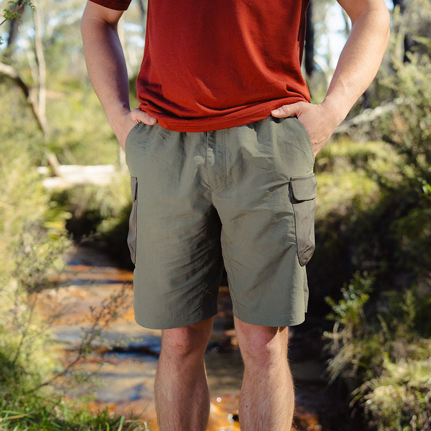 Person wearing a red shirt and green shorts standing in a natural setting with trees and a stream.
