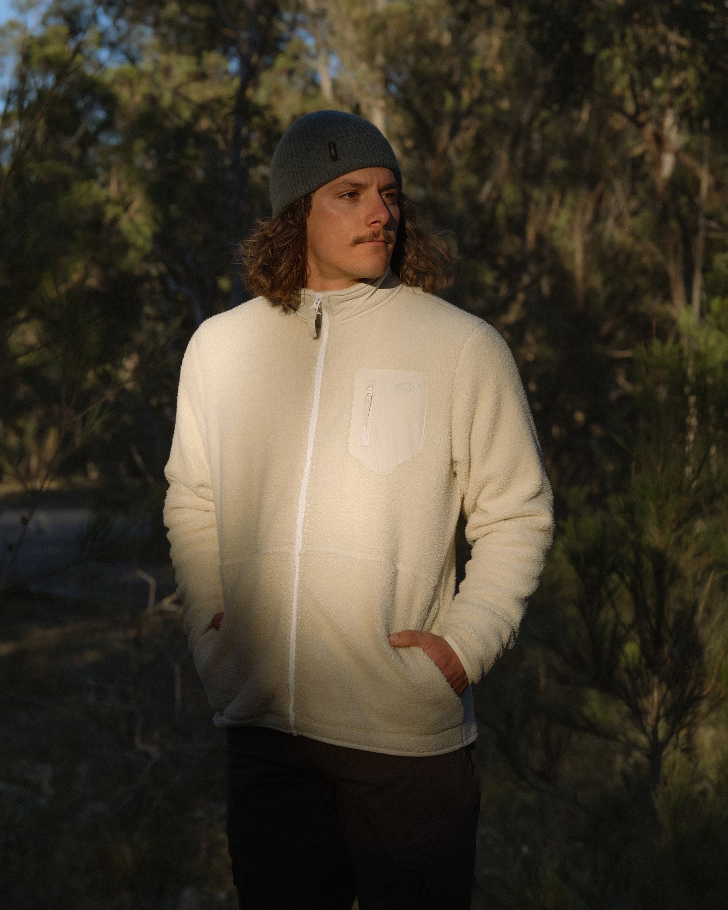Man wearing a cream Cosy Camp fleece jacket and grey beanie, standing outdoors in front of trees with hands in pockets.