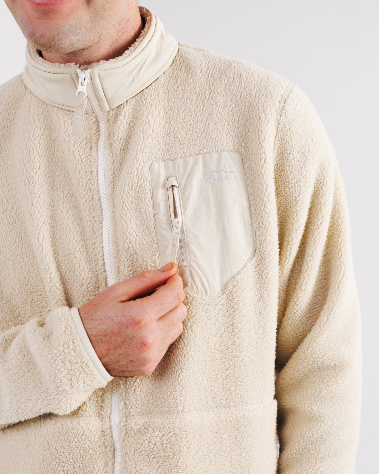Close-up of a cream sherpa fleece jacket's chest pocket with Zorali mountain logo, zipper open, model's hand reaching for it.