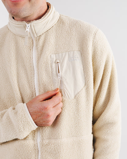 Close-up of a cream sherpa fleece jacket's chest pocket with Zorali mountain logo, zipper open, model's hand reaching for it.