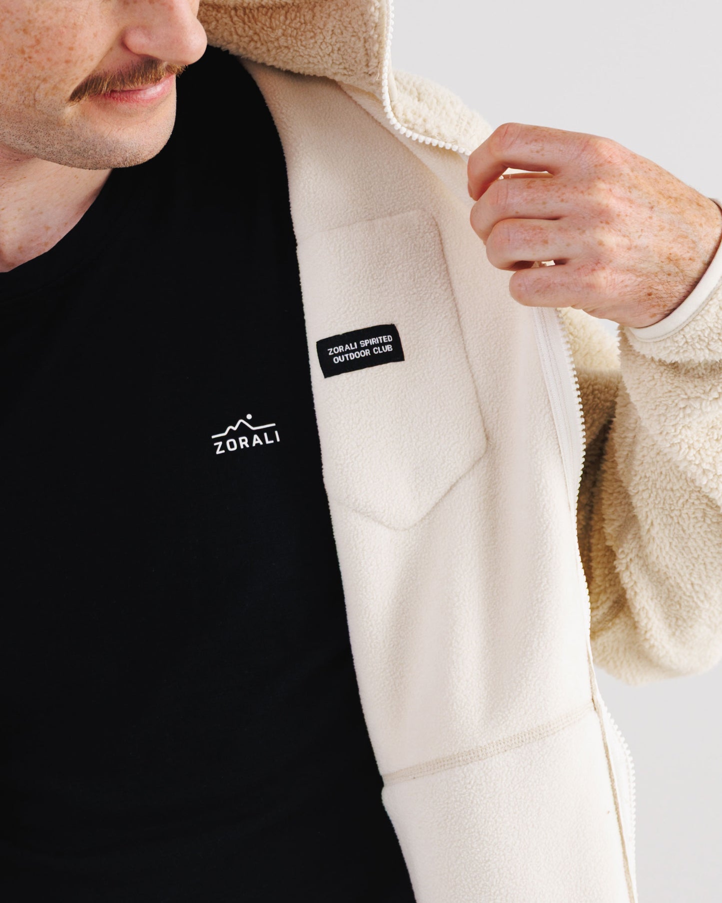 Close-up of a man opening his cream Cosy Camp fleece jacket to reveal the black interior lining and 'Zorali Spirited Outdoor Club' woven patch.