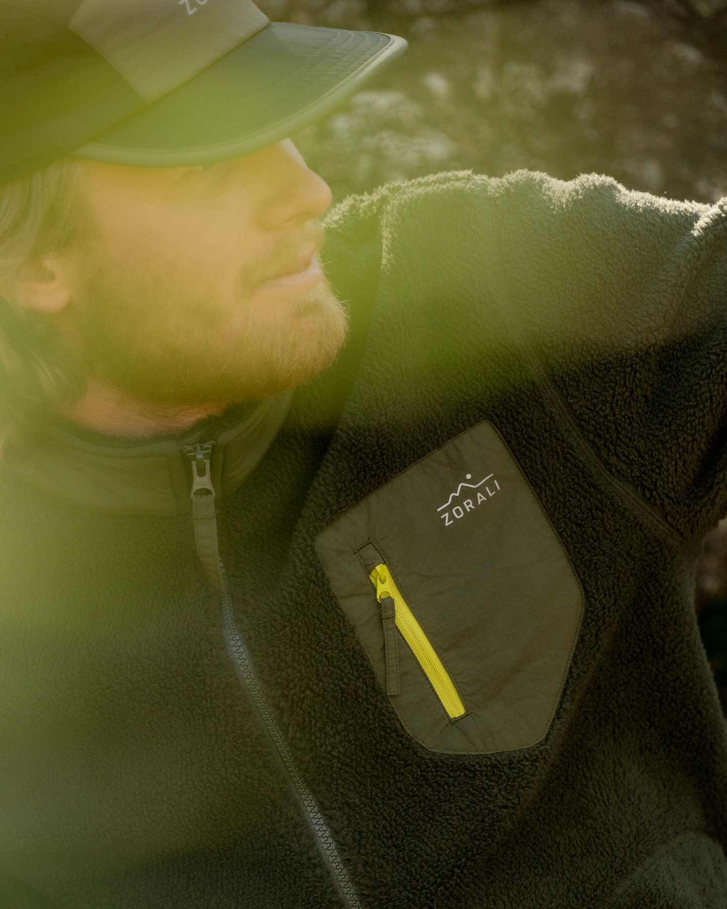 A man wearing a dark fleece jacket with a zippered chest pocket featuring a white Zorali logo and a bright yellow zipper pull, photographed outdoors with warm sunlight and lens flare.