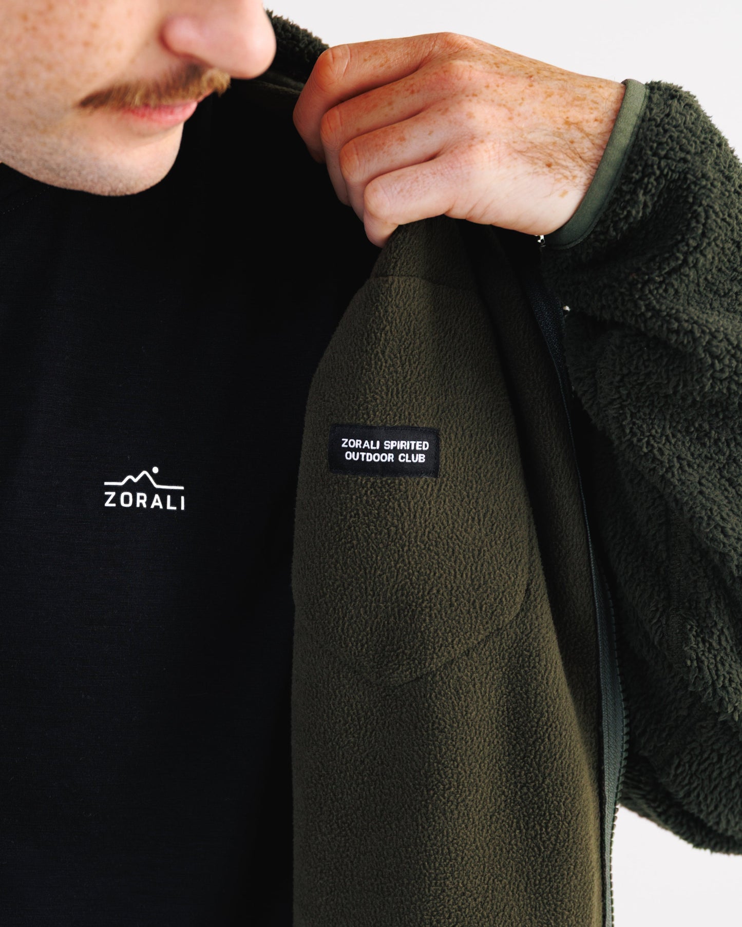 Close-up of a person wearing a dark olive green fleece jacket over a black shirt. The person is holding the collar of the jacket open, showing a black brand patch on the chest that reads 'ZORALI SPORTS OUTDOOR CLUB'.