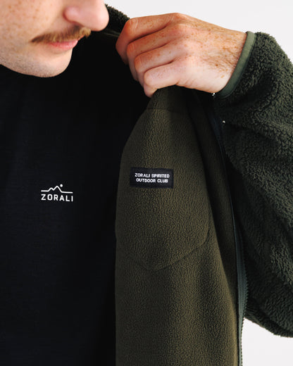 Close-up of a person wearing a dark olive green fleece jacket over a black shirt. The person is holding the collar of the jacket open, showing a black brand patch on the chest that reads 'ZORALI SPORTS OUTDOOR CLUB'.