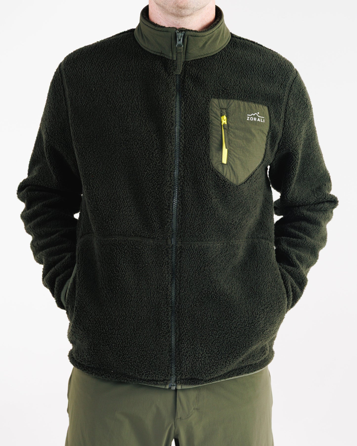 Man wearing a dark olive green sherpa fleece zip-up jacket, cropped view from chin to hip with hands in the pockets, showing the chest pocket with yellow zipper pull and Zorali logo.