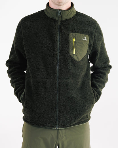 Man wearing a dark olive green sherpa fleece zip-up jacket, cropped view from chin to hip with hands in the pockets, showing the chest pocket with yellow zipper pull and Zorali logo.
