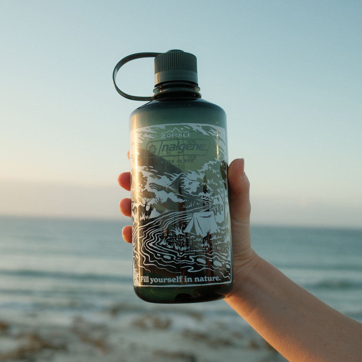Hand holding a water bottle with a scenic design against a beach backdrop
