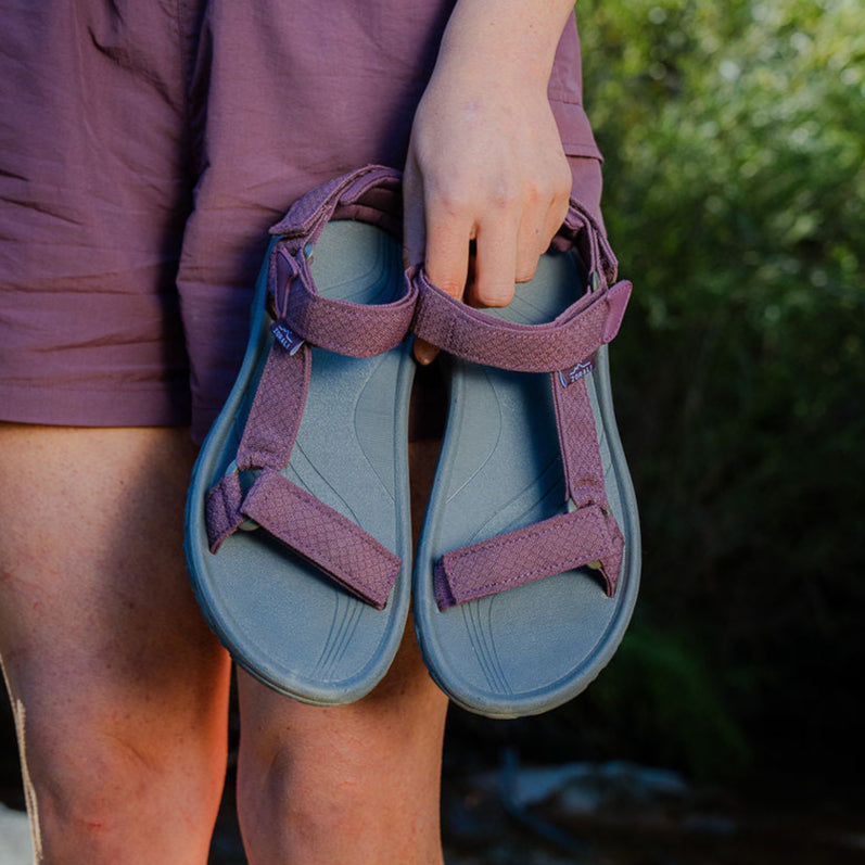 Person holding a pair of blue sandals with purple straps outdoors