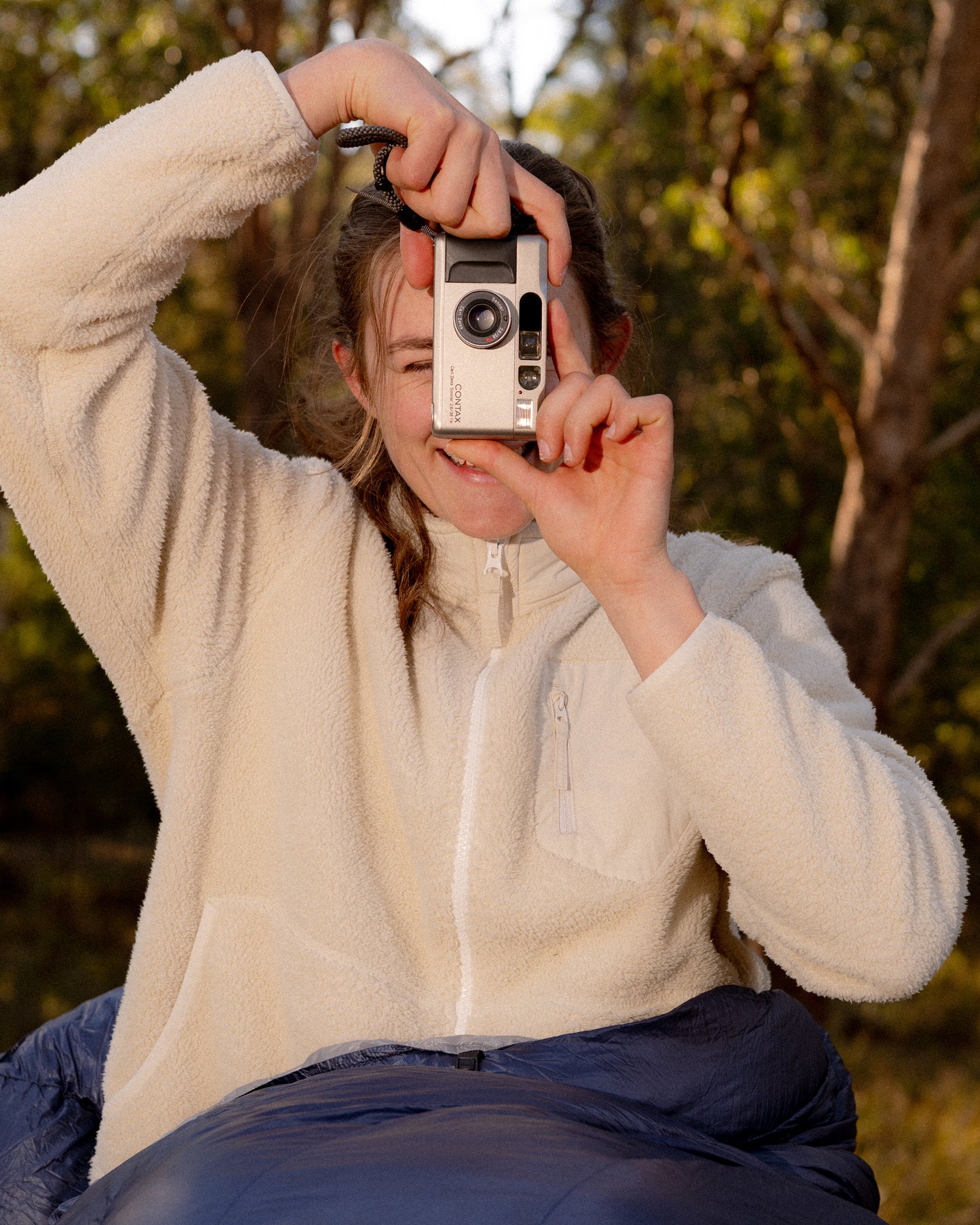 A person outdoors wearing a cream fleece zip-up jacket and dark blue pants, holding a retro-style silver camera in front of their face against a blurred forest background.