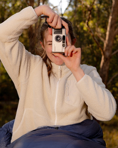 A person outdoors wearing a cream fleece zip-up jacket and dark blue pants, holding a retro-style silver camera in front of their face against a blurred forest background.