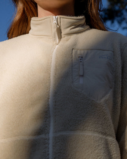 Close-up of a person wearing a cream fleece zip-up jacket outdoors, with sunlight casting shadows across the chest and a small visible ZORALI logo on the chest pocket.
