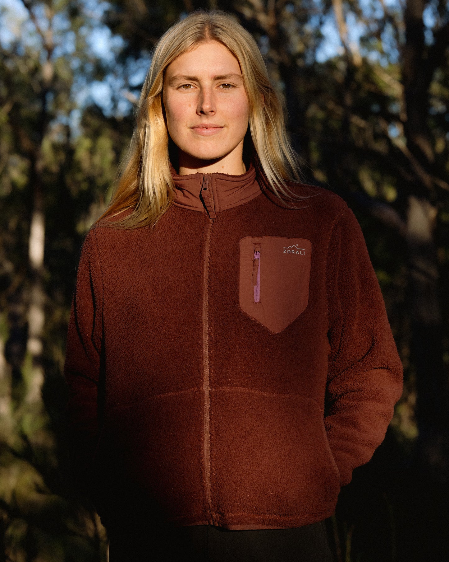 A woman with blonde hair wearing a brown fleece zip-up jacket with a chest pocket, standing outdoors against a dark, wooded background.