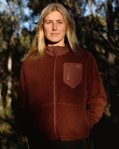 A woman with blonde hair wearing a brown fleece zip-up jacket with a chest pocket, standing outdoors against a dark, wooded background.