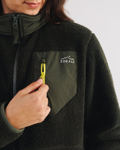 Close-up of a woman's hand unzipping the chest pocket on a dark green Cosy Camp fleece jacket, showing the Zorali logo.