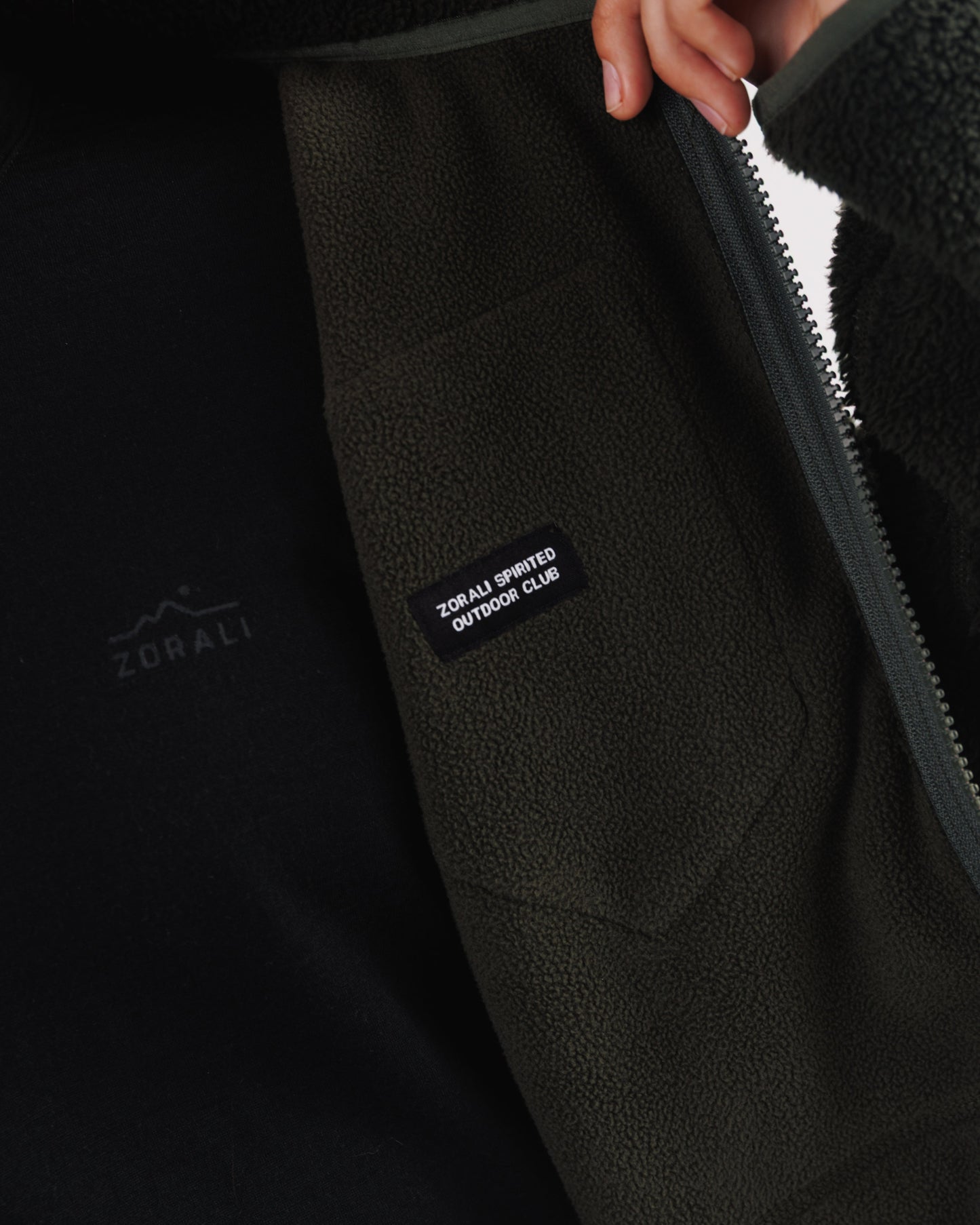Close-up of a hand pulling open the front zip on a dark green sherpa fleece jacket, revealing the 'Zorali Spirited Outdoor Club' patch inside.