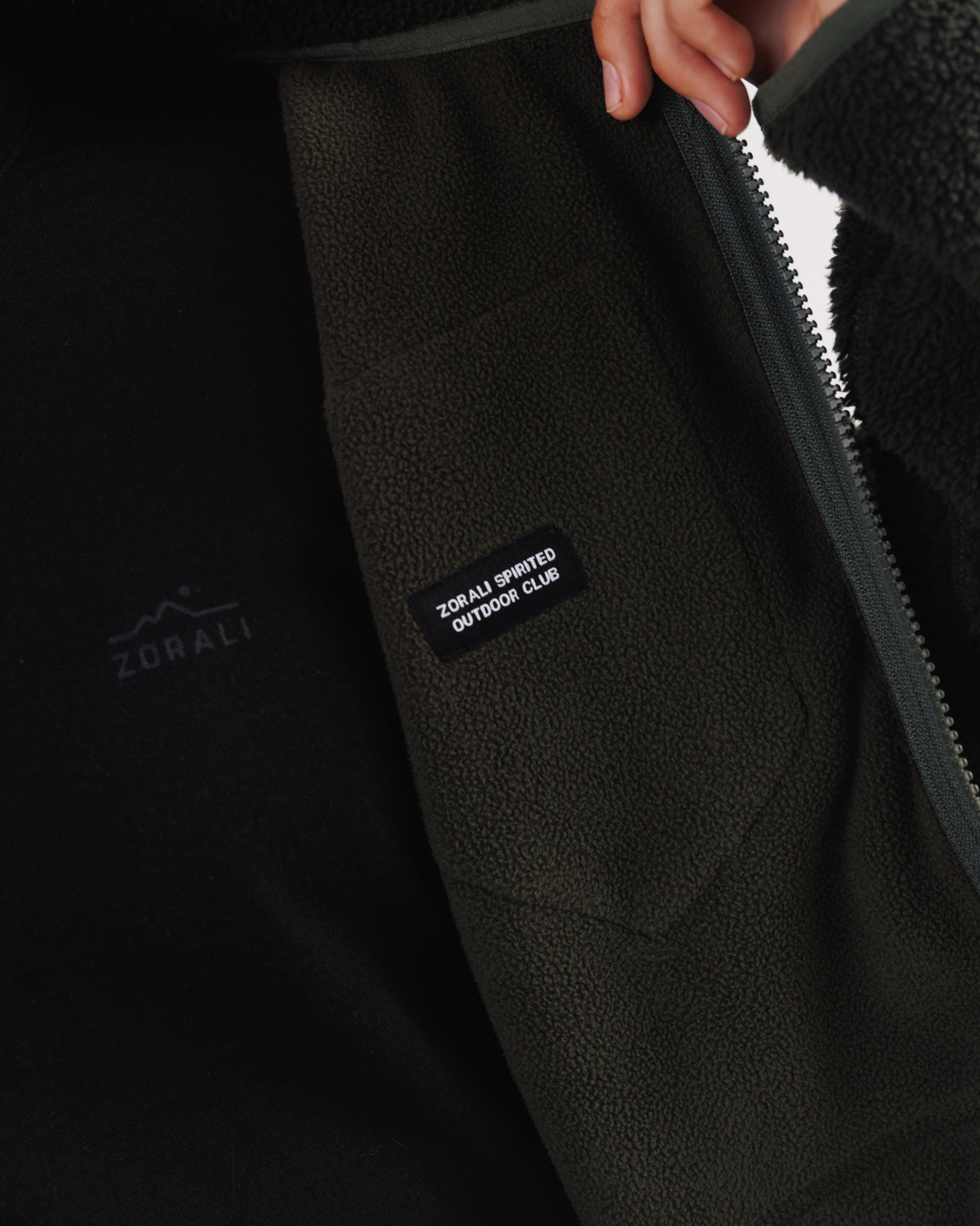 Close-up of a hand pulling open the front zip on a dark green sherpa fleece jacket, revealing the 'Zorali Spirited Outdoor Club' patch inside.
