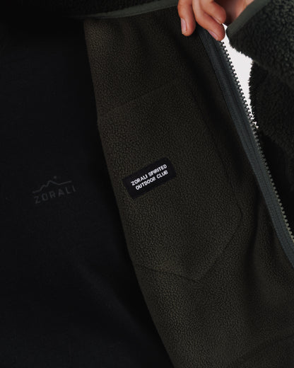 Close-up of a hand pulling open the front zip on a dark green sherpa fleece jacket, revealing the 'Zorali Spirited Outdoor Club' patch inside.