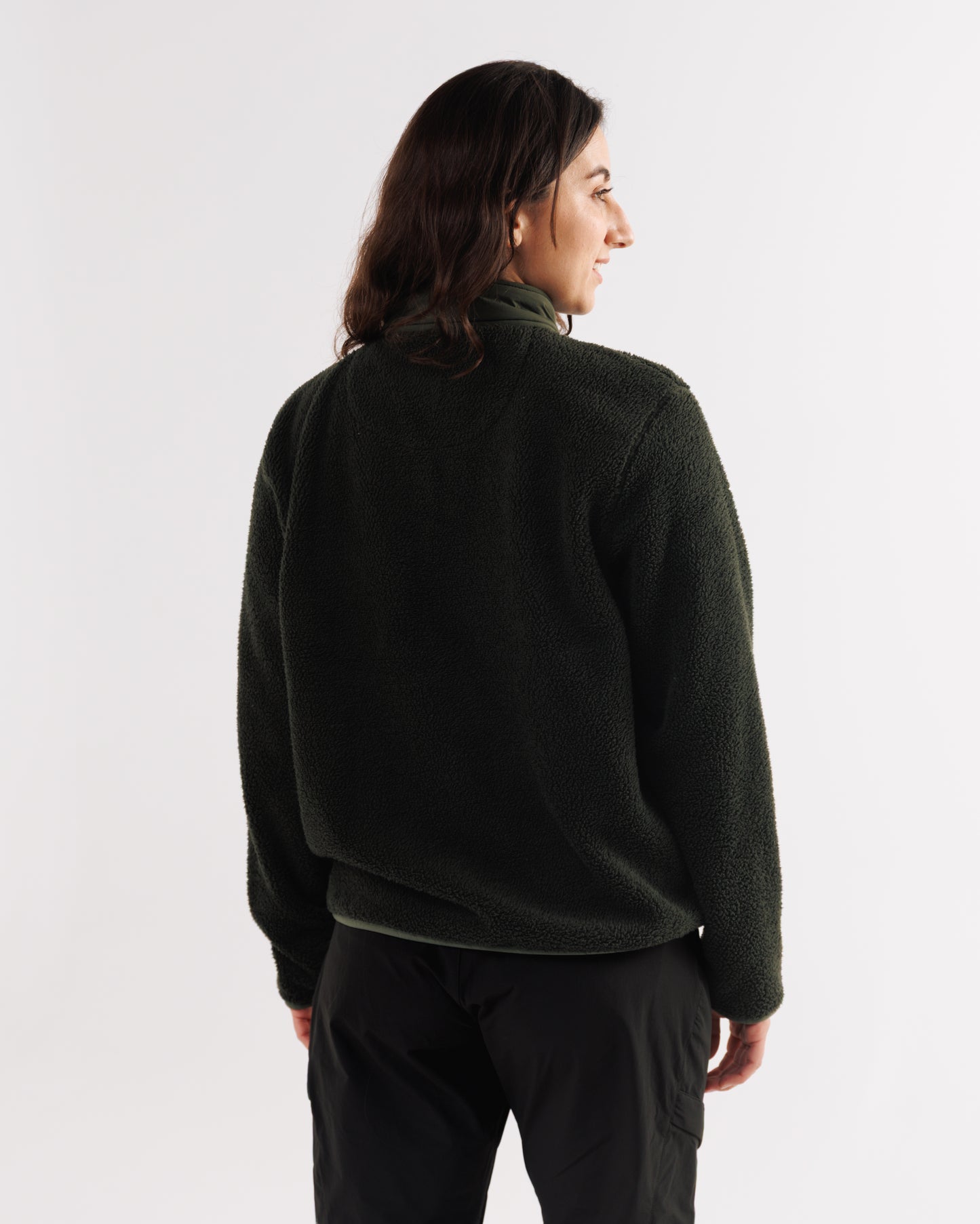 Back view of a woman wearing a dark green sherpa fleece jacket, smiling over her shoulder.