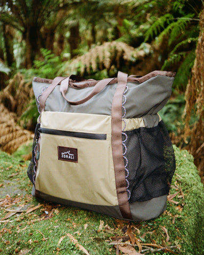 A large tote bag with a beige and gray body, brown straps, and a black mesh side pocket, resting on a mossy log in a lush green forest.