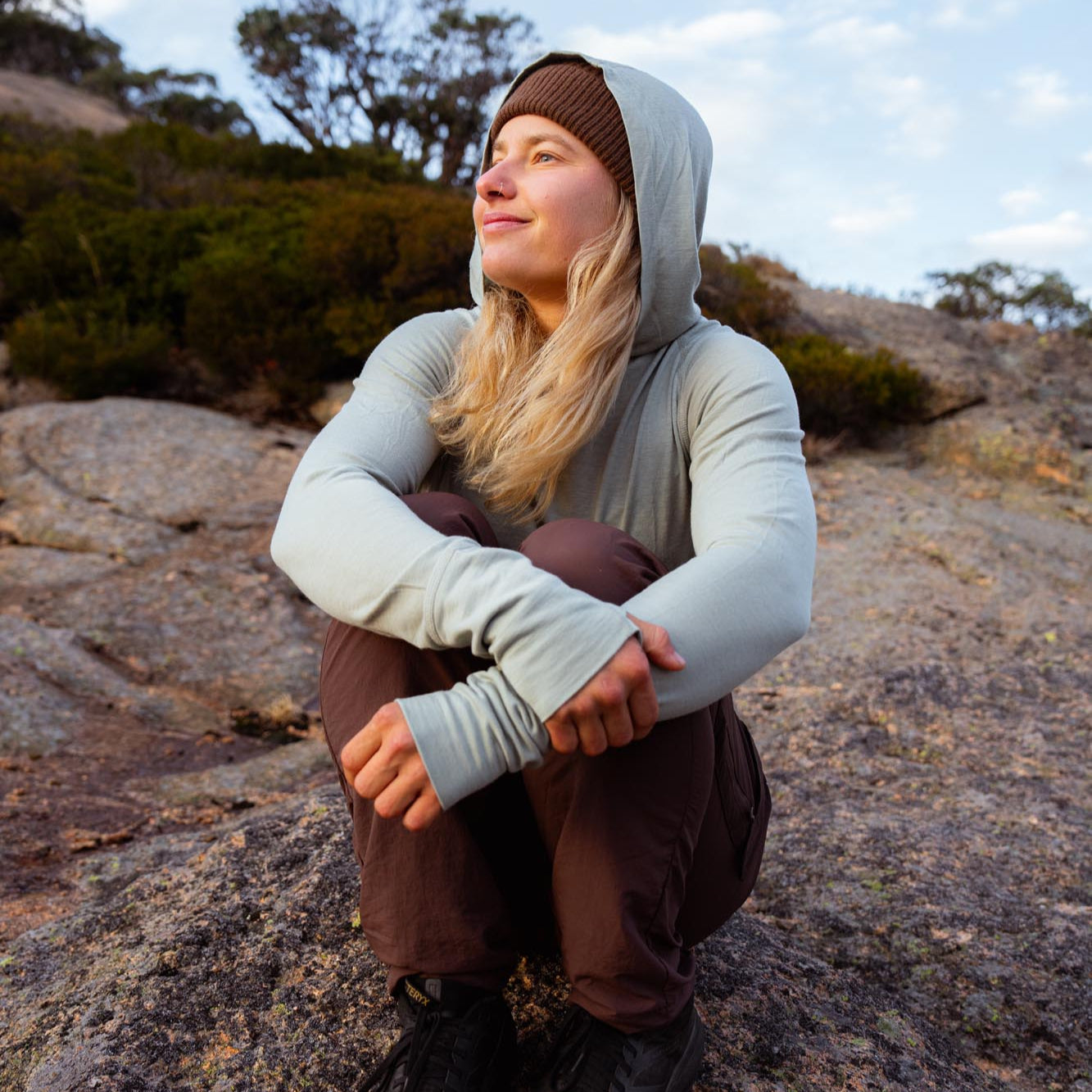 Person sitting on a rock outdoors wearing a hooded merino longsleeve top and brown pants.