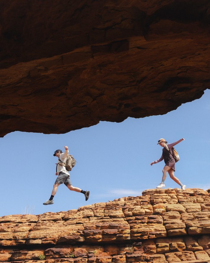 Two hikers leaping across a sandstone ridge under a rock arch at Kings Canyon